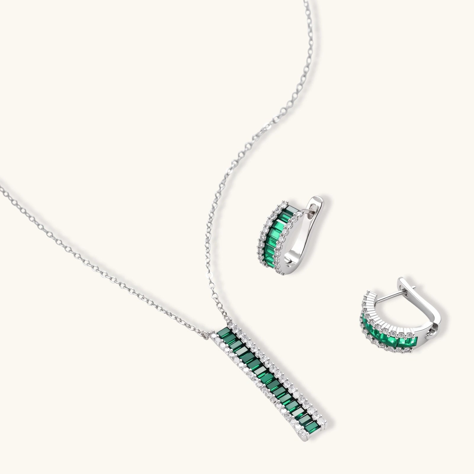 White gold plated jewelry set with emerald green rectangular stones and clear crystals in geometric rows on white background.