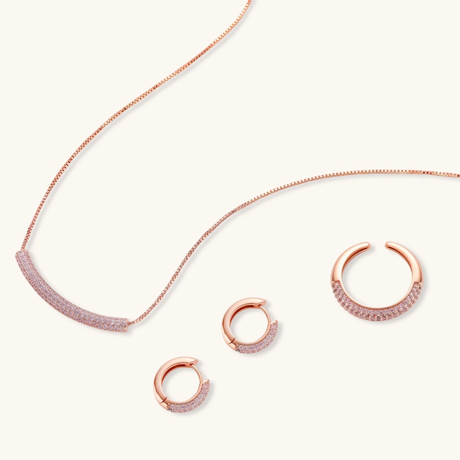 Rose gold pavé zirconia bar necklace, hoop earrings, and open ring set with pink stones on cream background.