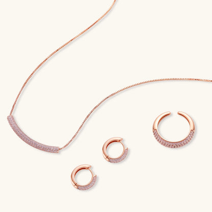 Rose gold pavé zirconia bar necklace, hoop earrings, and open ring set with pink stones on cream background.