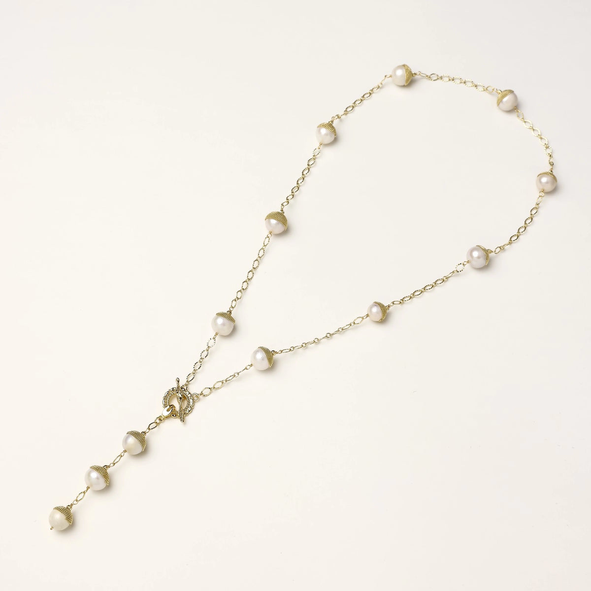 Gold-plated lariat necklace with baroque pearls and textured caps on cream background, showing adjustable Y-shaped design.