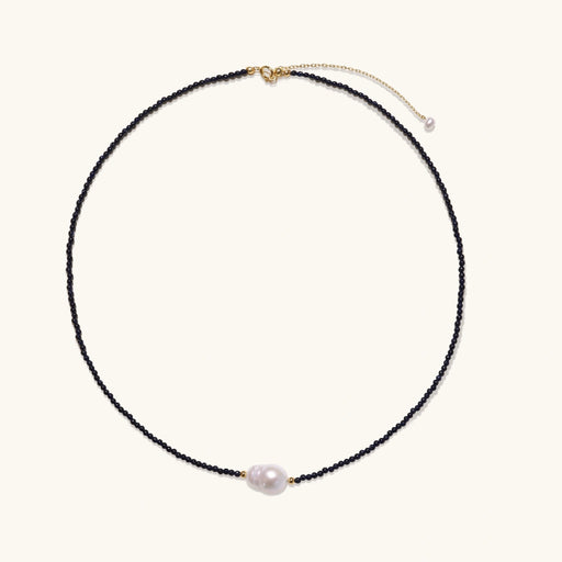 Delicate necklace with black blue sandstone beads and baroque pearl center on white background with gold accents.