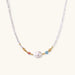 Delicate white beaded necklace with baroque pearl center, gold accents, and coral and aqua gemstones on cream background.
