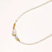 Delicate white beaded necklace with baroque pearl center, gold accents, and coral and aqua gemstones on cream background.
