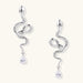Silver snake-shaped drop earrings with crystal accents and teardrop stone dangles on cream background.