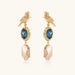 Gold-plated bird stud earrings with iridescent abalone shell medallions and champagne crystal drops on cream background