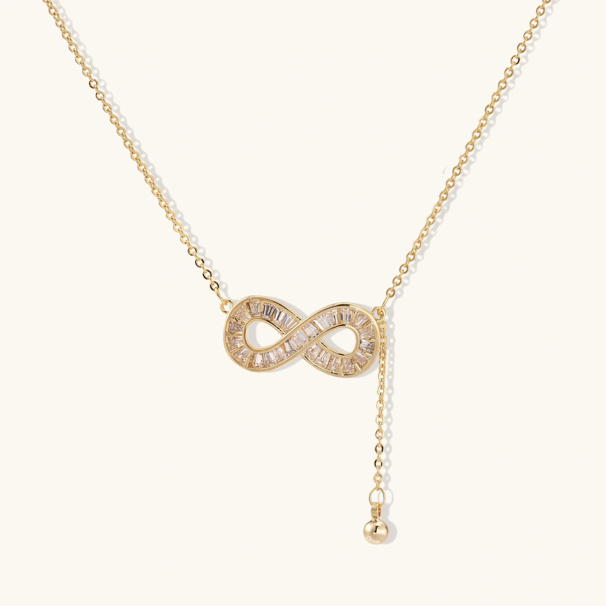 Gold infinity pendant with baguette crystals on adjustable chain against cream background.