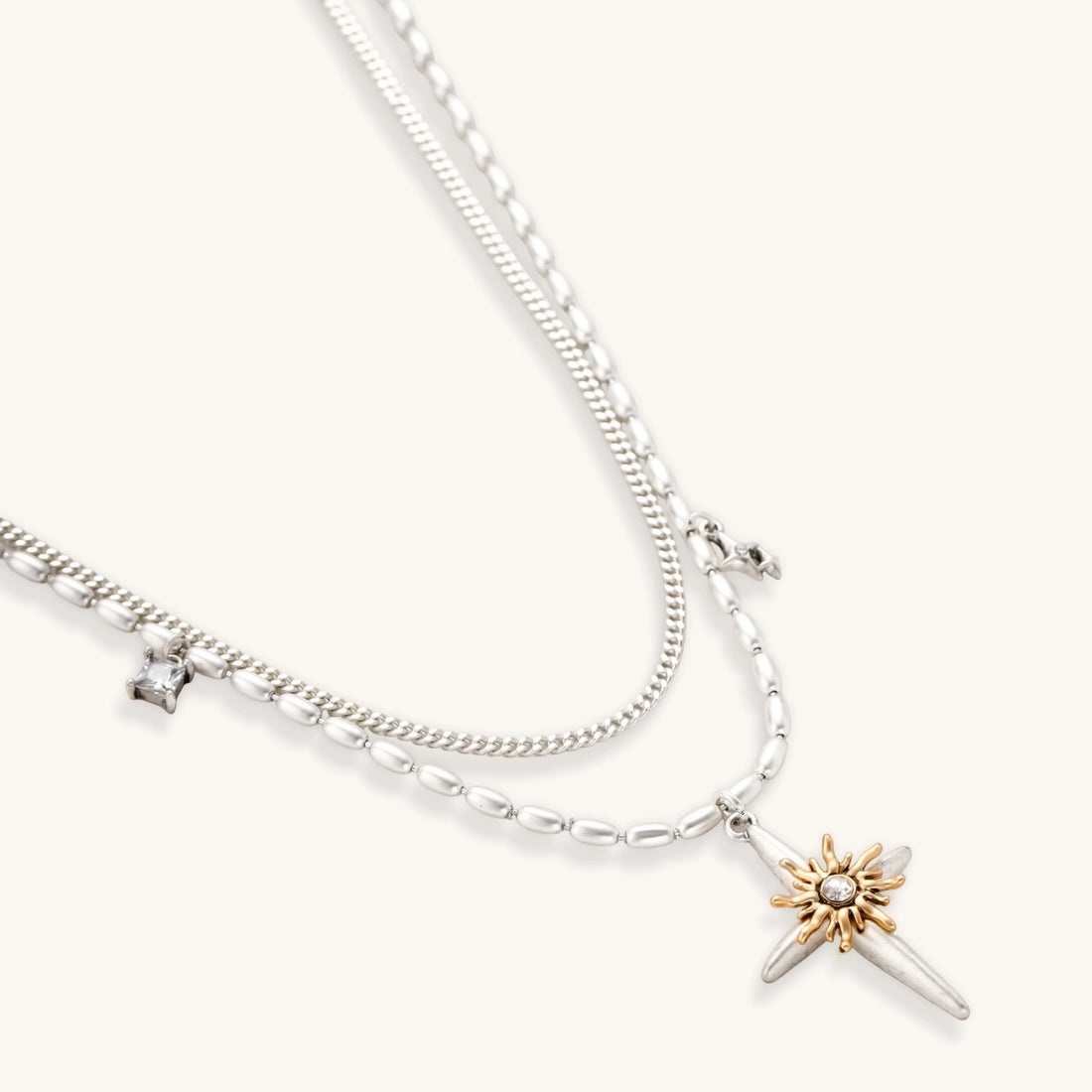 Layered silver-toned necklace set with cross pendant and gold sun charm detail, matching drop earrings on white background.