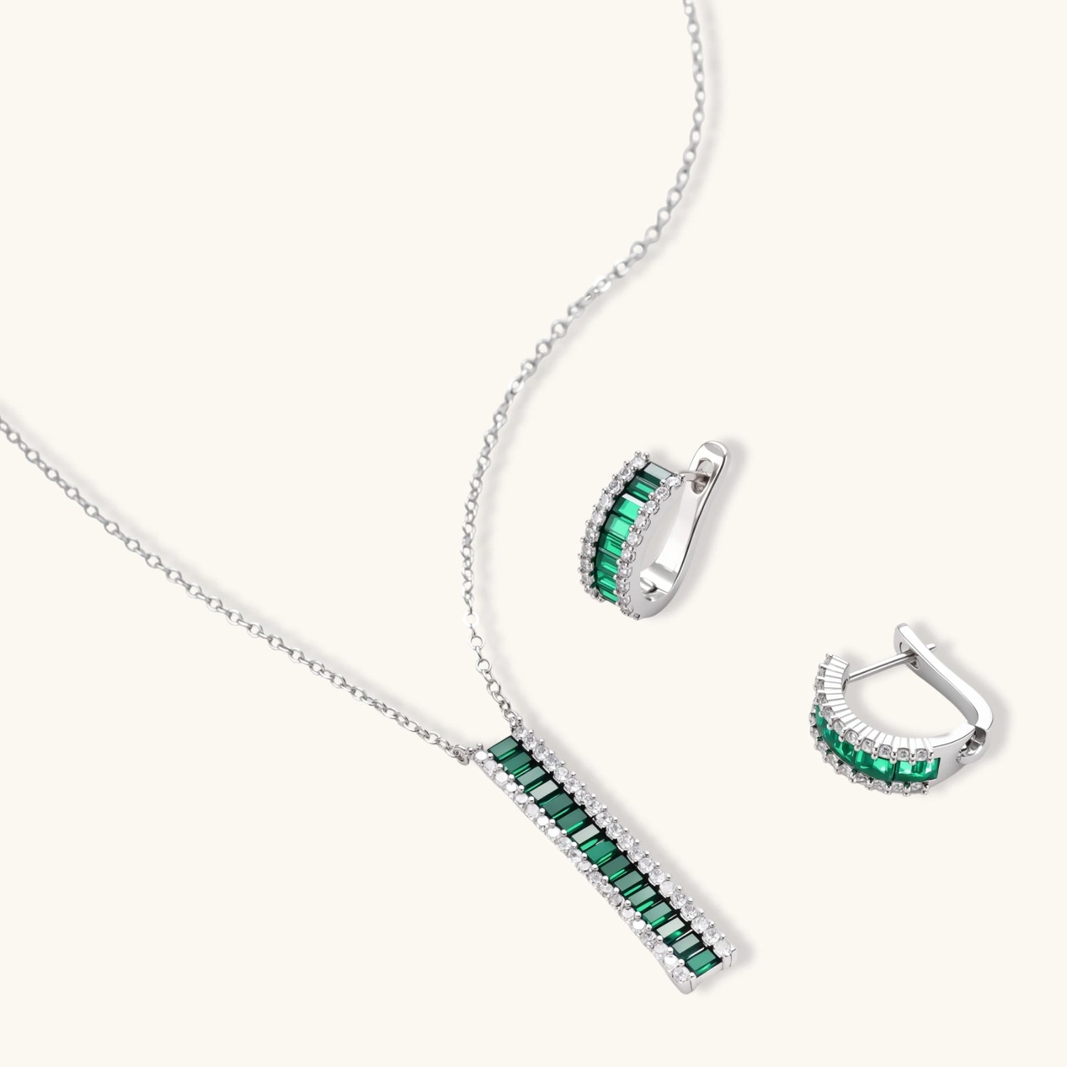 White gold plated jewelry set with emerald green rectangular stones and clear crystals in geometric rows on white background.